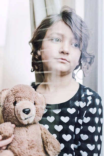 istock Pensive child looking through a window with her teddy bear 169993729