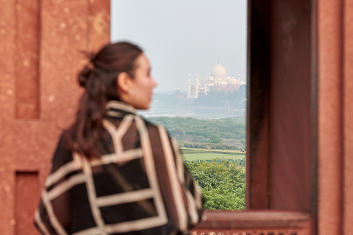 Rear view east asian woman in black dress look at Taj Mahal through window opening, back view to asian girl admires beautiful indian landscape, female tourist covered white scarf rear view in India