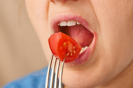 Close up happy Caucasian woman brings a slice of fresh tomato on a fork to her open mouth. Three-quarter front view. Low angle view.