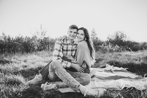 Couple sitting on blanket, hugging in grass in field at sunset. Happy young woman and man spending time together in nature. Concept family holiday. Female embrace male on picnic. Black and white photo