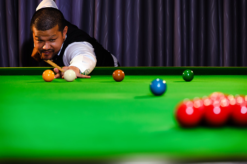 Asian snooker player while aiming to white ball shoot to hit the red ball group set in game on snooker table.