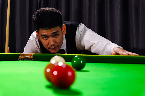 Asian snooker player while aiming to white ball shoot to hit the Snooker ball in game on snooker table.