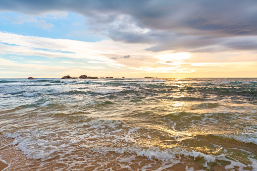 A beautiful view of the sun setting over the ocean and the rocks on a wet sandy beach. The sky is a blend of orange, yellow and blue colors with clouds scattered across it. The sun is partially hidden by the clouds and creates a bright spot on the horizon. The ocean is a mix of blue and green colors with white foam on the waves. The waves are breaking on the shore and create a contrast with the rocks in the distance. The sand is a light brown color and is wet from the waves.
