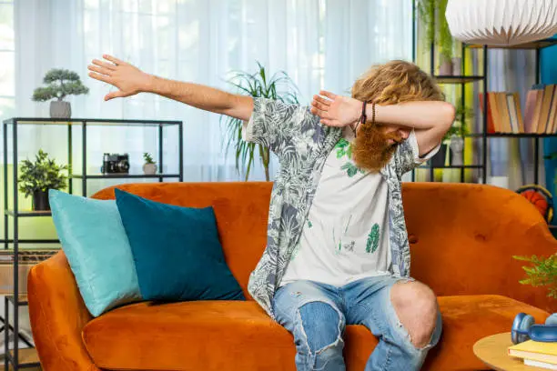 Photo of Happy man having fun, dancing, dabbing raising hands, making dub dance gesture at home room on sofa