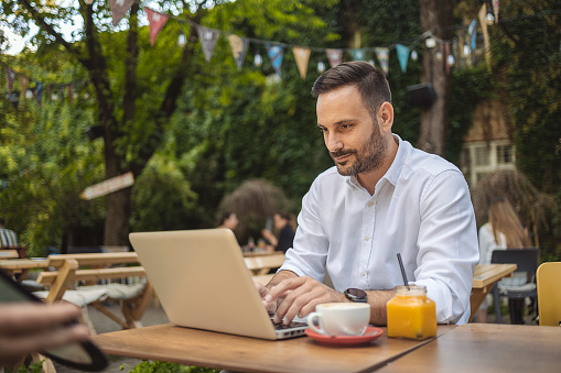 Young Stylish Businessman in White Shirt Working on Laptop Computer While Sitting at a Cafe Outdoors