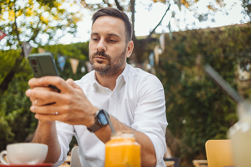 Man in White Shirt Using Smart Phone While Sitting at a Cafe Outdoors