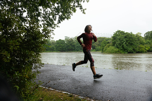 A shot of a female runner taking part in a community fun run in the rain in Leazes Park in Newcastle upon Tyne, North East England. She is smiling as she runs, behind her is a lake and a tree area. The race is open to people of all ages and abilities and is also dog friendly. \n\nThese files have similar videos/images available.