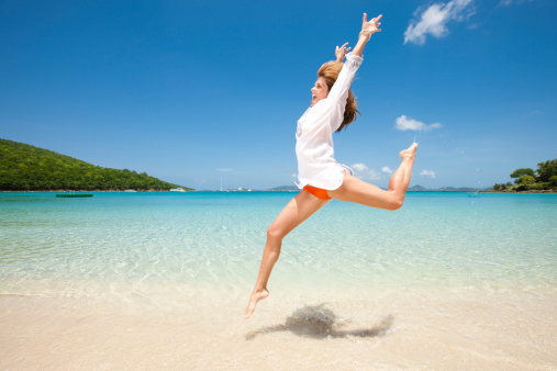 A carefree mid-adult woman celebrates her freedom by jumping for joy like a ballerina on the beach while on vacation on an idylic tropical island.