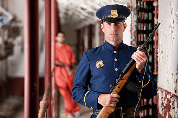 A ruggedly handsome corrections officer, holding a rifle, guards a delapidated cell block and the prisoners within.