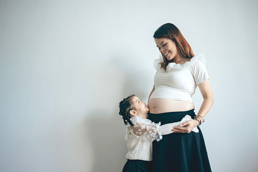 A young Asian girl is holding her mother's belly, feeling the baby's movements and heartbeat, showcasing the love and care within the family, as well as the anticipation for the arrival of a new member.
