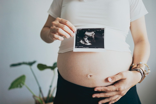 In a close-up shot, a pregnant woman gently touches her belly while holding an ultrasound scan photo against a white background. She is preparing to welcome a new family member and is filled with anticipation for the arrival of new life. This scene embodies the concepts of love and care, as well as the responsibilities of parenthood.