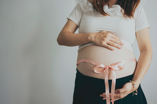 In a close-up shot, an Asian pregnant woman stands against a white background with a ribbon tied around her belly. This symbolizes the anticipation for the new baby, much like a gift within the family.