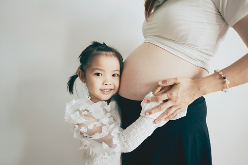 A close-up shot captures the expression of an Asian young girl as she eagerly anticipates her mother's unborn baby. She embraces her mother, showcasing the love within the family and the sibling bond, all while gazing directly into the camera.