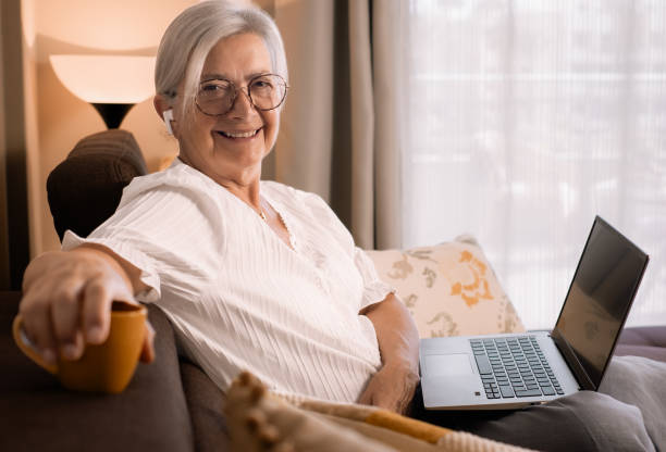 istock Attractive senior white haired woman sitting on home sofa using laptop looking at camera smiling. Elderly relaxed lady watching movie or social wearing earphones 1700104758