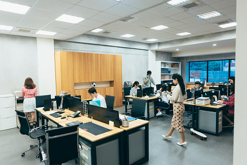 A busy office, where employees are engrossed in various tasks. Phones are constantly ringing, and stacks of documents cover the desks. Computer screens display data and charts, while various office supplies are scattered across workstations. Staff members are bustling around, creating a dynamic and active atmosphere in the office.