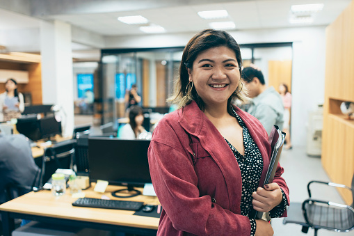 An Asian female businesswoman is looking at the camera with a busy office background. She may be standing in the office, focused on interacting with the camera, while the office background reflects a bustling work atmosphere, filled with the activities of employees and office tasks.