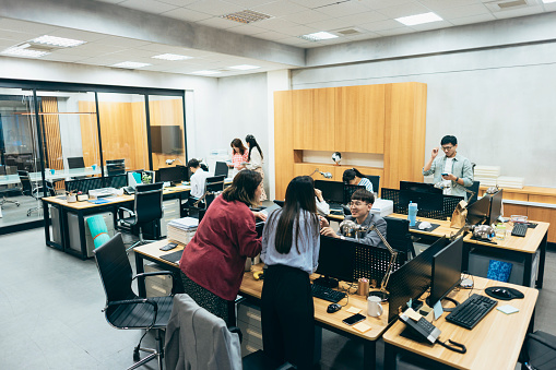 A busy office, where employees are engrossed in various tasks. Phones are constantly ringing, and stacks of documents cover the desks. Computer screens display data and charts, while various office supplies are scattered across workstations. Staff members are bustling around, creating a dynamic and active atmosphere in the office.