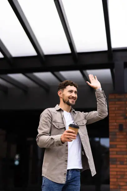 Attractive man standing outside waving hand, saying goodbye to his groupmates, coffee-to-go