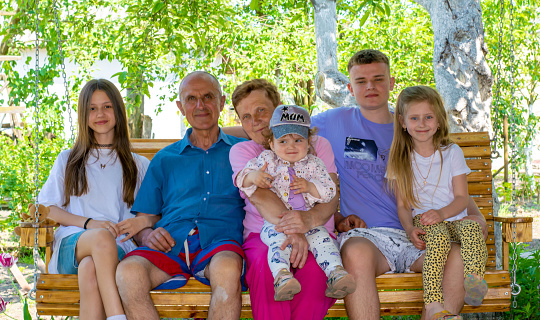 A family, grandparents, and their grandchildren sit on a swing outside their house.