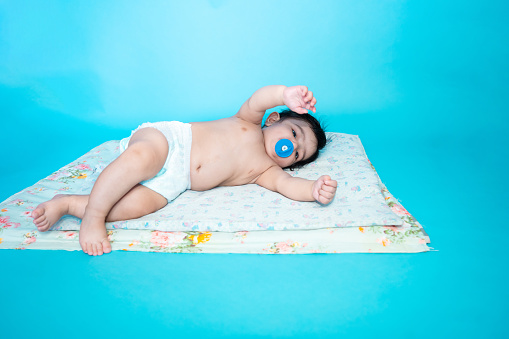 Small six months old cute indian baby boy wearing diaper lying on floor with nipple soother or pacifier isolated over blue studio background, copy space.