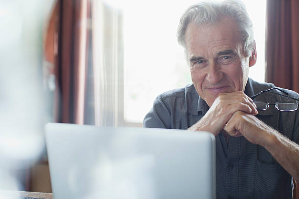 retrato de homem senior sorridente segurando óculos e usando computador portátil - senior adult happiness computer looking at camera imagens e fotografias de stock