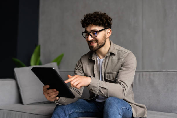 istock Handsome young man is using a digital tablet and smiling while resting on couch at home 1700156997