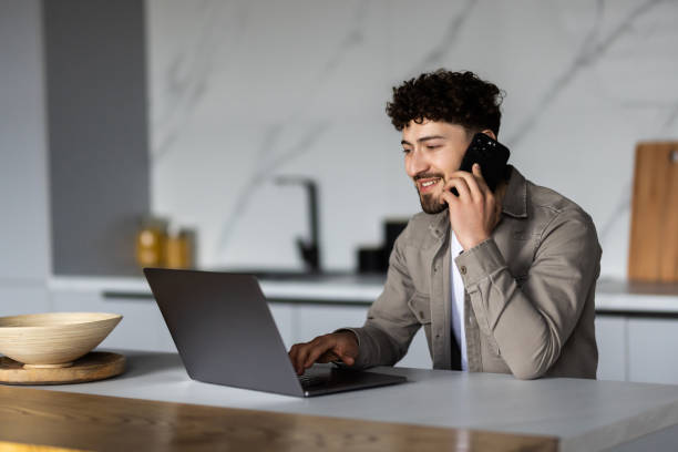 istock Young man freelancer working remotely at home, looking at laptop and consulting client via cellphone, sitting at kitchen 1700181370