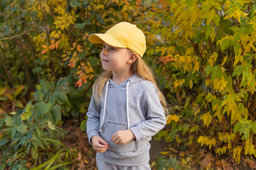 Portrait of a cute smiling little girl with blonde hair in a yellow cap on a background of orange and yellow leaves in an autumnal sunny day.