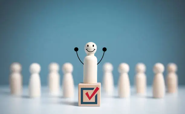 Photo of Raise hand wooden figure stands above a wooden cube with vote symbol among another. open-mindedness or public hearing, Elections concept. Volunteers, parties, candidates, constituency electorates.