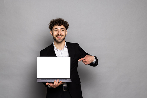 Optimistic man pointing finger at laptop with blank display, showing empty screen, bragging with internet advertisement, wearing official style suit. Indoor studio shot isolated on gray background.