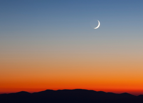 Crescent Moon rising over mountains and orange sunset sky  