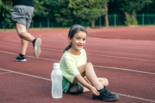 A cute sporty little girl with bottle of water sitting on running track at the stadium.