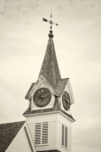An old, weather-beaten New England wooden church steeple with broken shingles and peeling paint - but with still complete, though weathered and damaged, wooden clocks on all sides. Sepia monotone with slight vignette added.