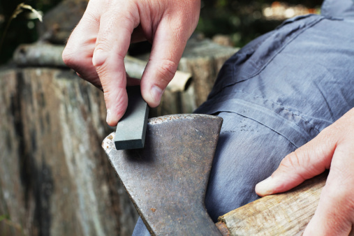 A man is sharpening an old, rusty axe manually using a whetstone.