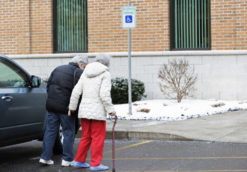 An elderly woman - supported by her cane on one side and her caregiver daughter on the other - is walking toward a doctor's office building for an appointment. It's cold and slippery in the winter snow and they are stepping cautiously through the scattered rock salt in the parking lot.