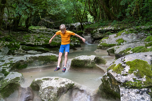 Active boy jumps from rock to rock on a calm mountain river hidden in a dense forest