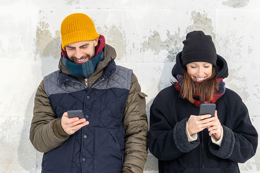 Smiling couple using smartphones on wall background outside. Laughing man and woman standing by white wall holding mobile phones. Positive people man and woman using cellphones on gray wall.