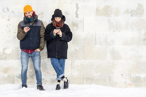 Laughing couple standing by white wall holding mobile phones. Smiling man and woman using smartphones on wall background outside. Positive people man and woman using cellphones on gray wall.