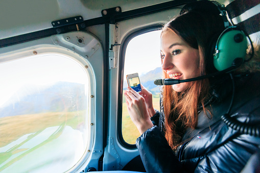 young woman enjoying a helicopter ride and taking photos of the landscape with her smartphone.
