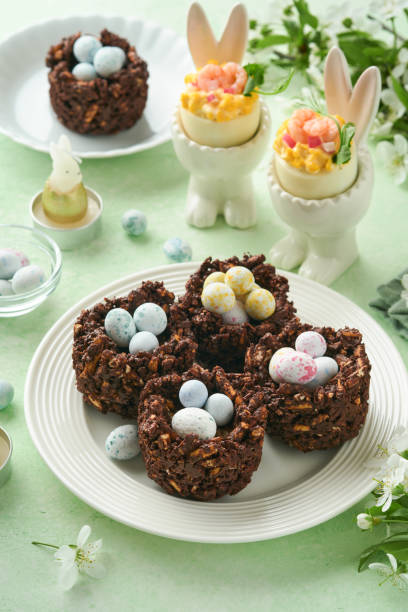 istock Easter chocolate nest cake with mini chocolate candy eggs with blossoming cherry or apple flowers on green background table. Creative recipe for Easter table with holiday decorations. Top view. 1700292879