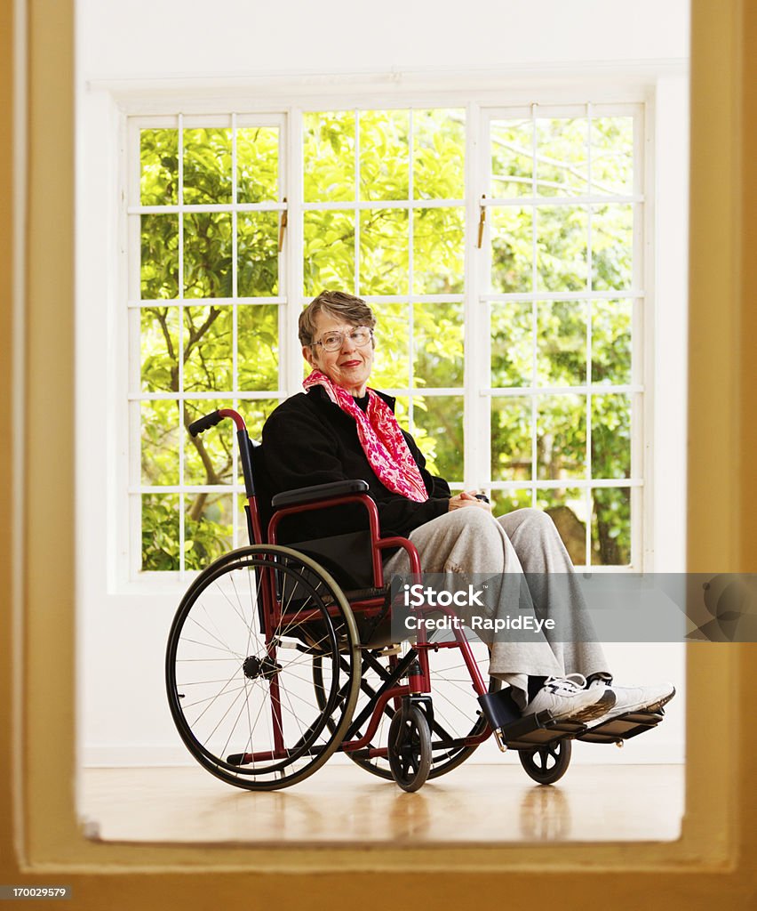 Edad mujer sentada en la silla de ruedas, enmarcado por una puerta, sonrisas suavemente - Foto de stock de 60-69 años libre de derechos