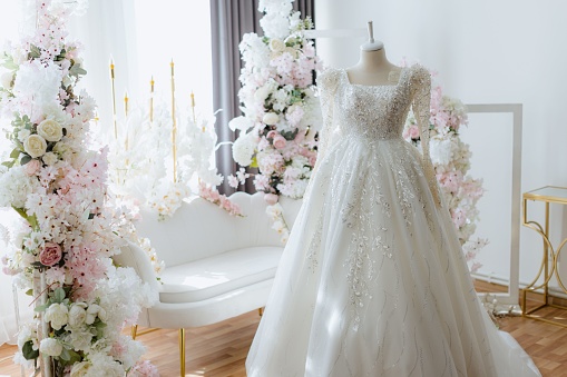 A beautiful white wedding dress is on display in a pristine white room decorated with vibrant flowers