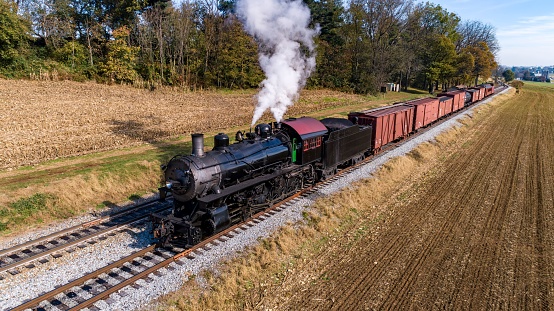 Steam train driving in the countryside with smoke coming from the chimney. The black and red locomotive is pulling passenger railroad car with tourists.