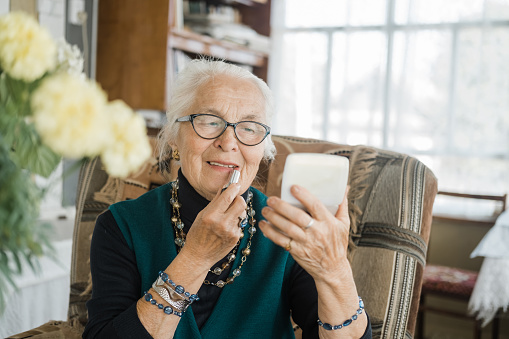 An elderly elegant woman with glasses with gray hair looks in the mirror and paints her lips with lipstick