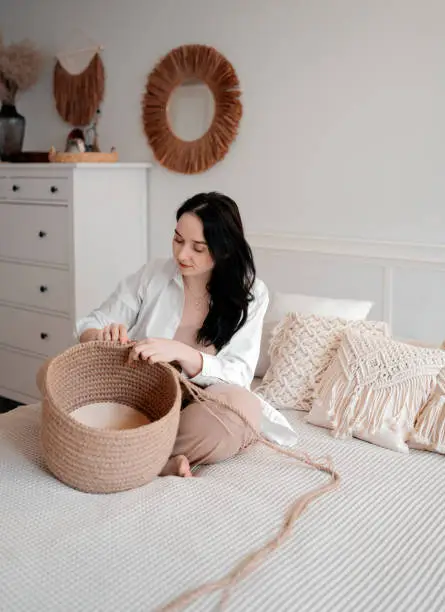 A girl with dark hair is sitting on the bed and knitting a basket of jute in a bright bedroom.