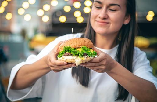 An attractive young woman in hands holds a burger, eating fast food in street cafe on a blurred background.