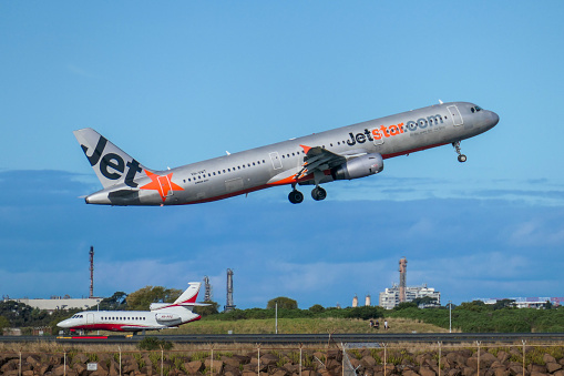 A Jetstar Airbus A321-231, registration VH-VWT, taking off to the south at Sydney Kingsford-Smith Airport and heading to Cairns as flight JQ958.  She is passing over a Dassault Falcon 900C, registration VH-PPD, just arrived from Wellington, New Zealand.  A small group of people is visible on the opposite grass bank.  This image was taken from Botany Bay, Kyeemagh on a sunny afternoon on 23 September 2023.