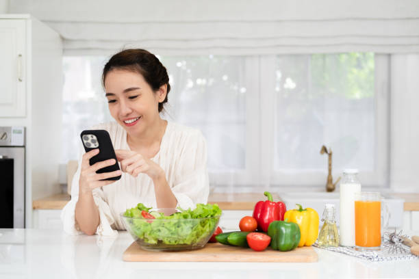 istock Smiling young woman using cellphone while cooking dinner in kitchen at home, cheerful casual female leaning on table, reading text messages on her cell phone and smiling, free copy space 1700337690