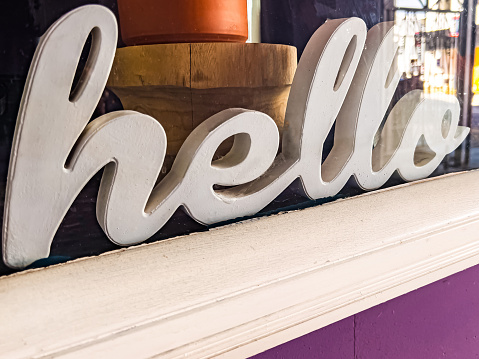 The word hello is written in cursive and is in a local small business storefront window. The dominate color is white with a touch of purple. The image is slanted.