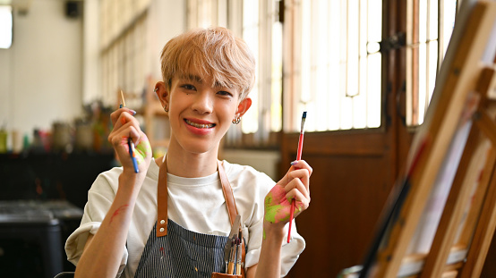 Portrait image of an attractive and smiling Asian male teenage artist with colored hair holding a paintbrush and pencil.